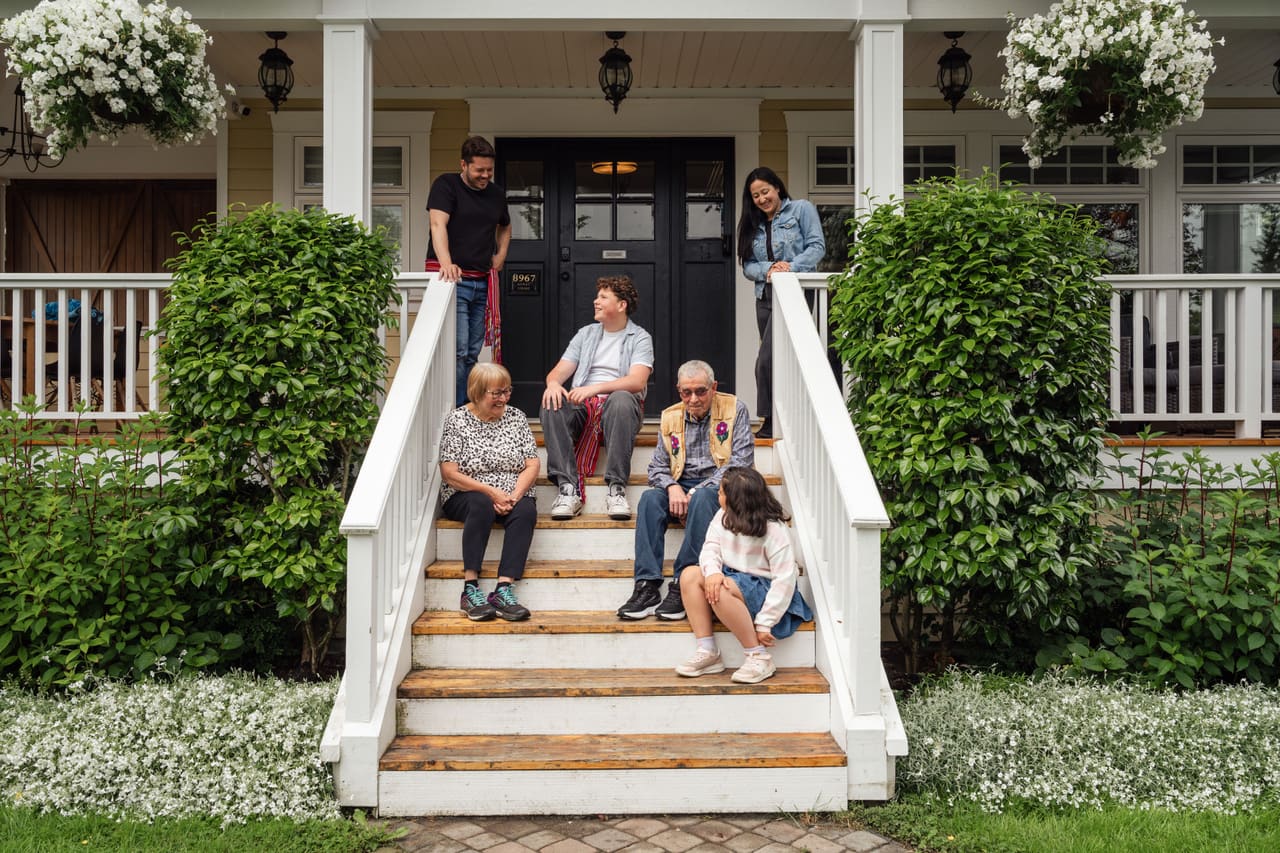 A multigenerational family of six gathered on the front steps and porch of a house, smiling and talking together.