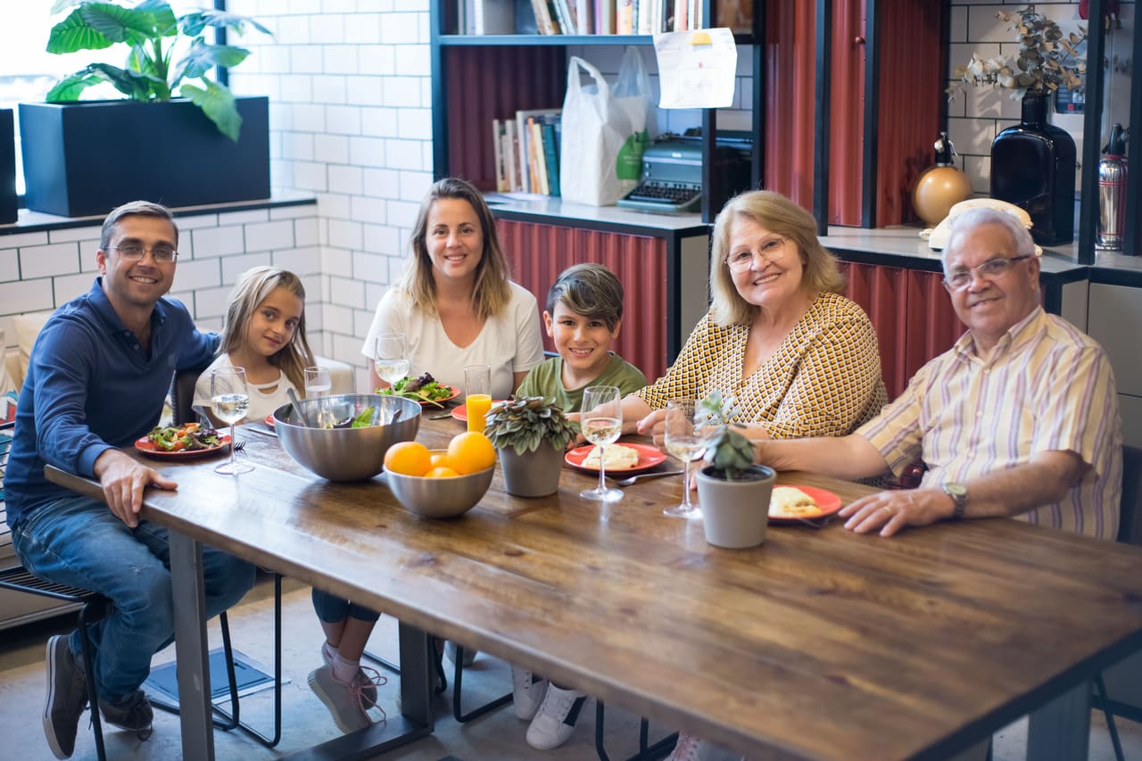 A multigenerational family of six seated together at a table with a meal, smiling at the camera.