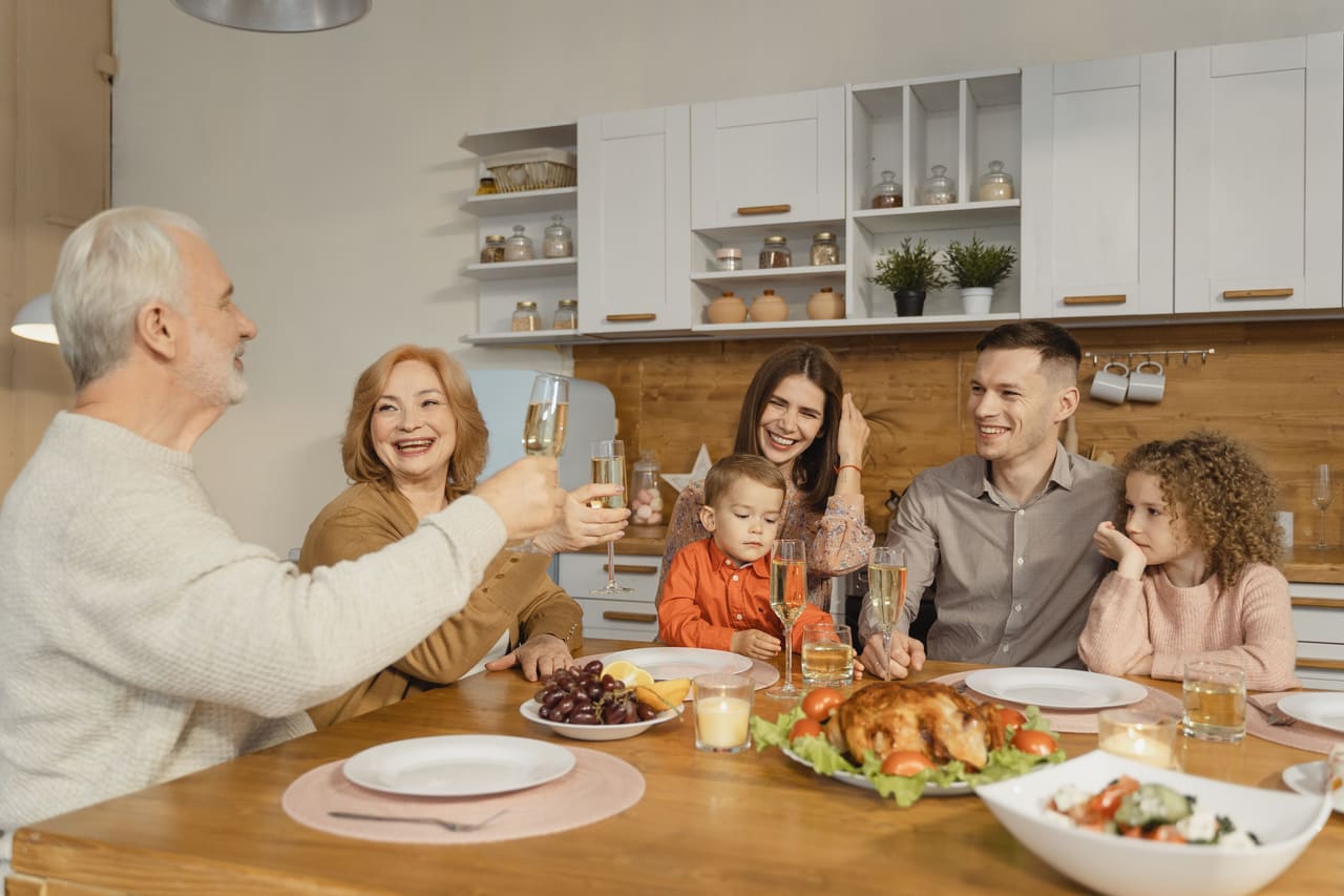 Three generations of a family sitting around a wooden table set with a roast dinner, raising champagne glasses together and laughing.