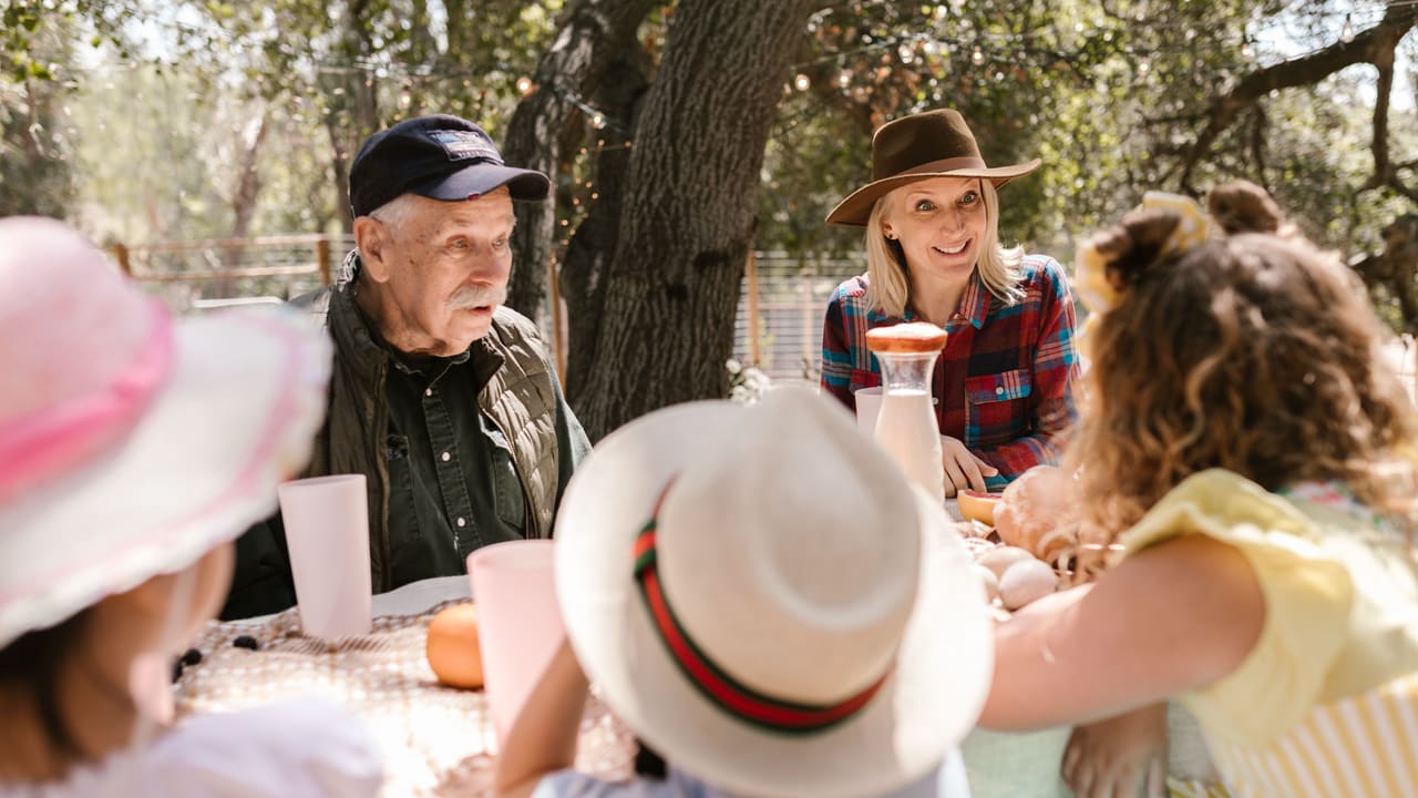 A multigenerational family wearing sun hats, gathered around an outdoor table set with bread and fruit under trees.